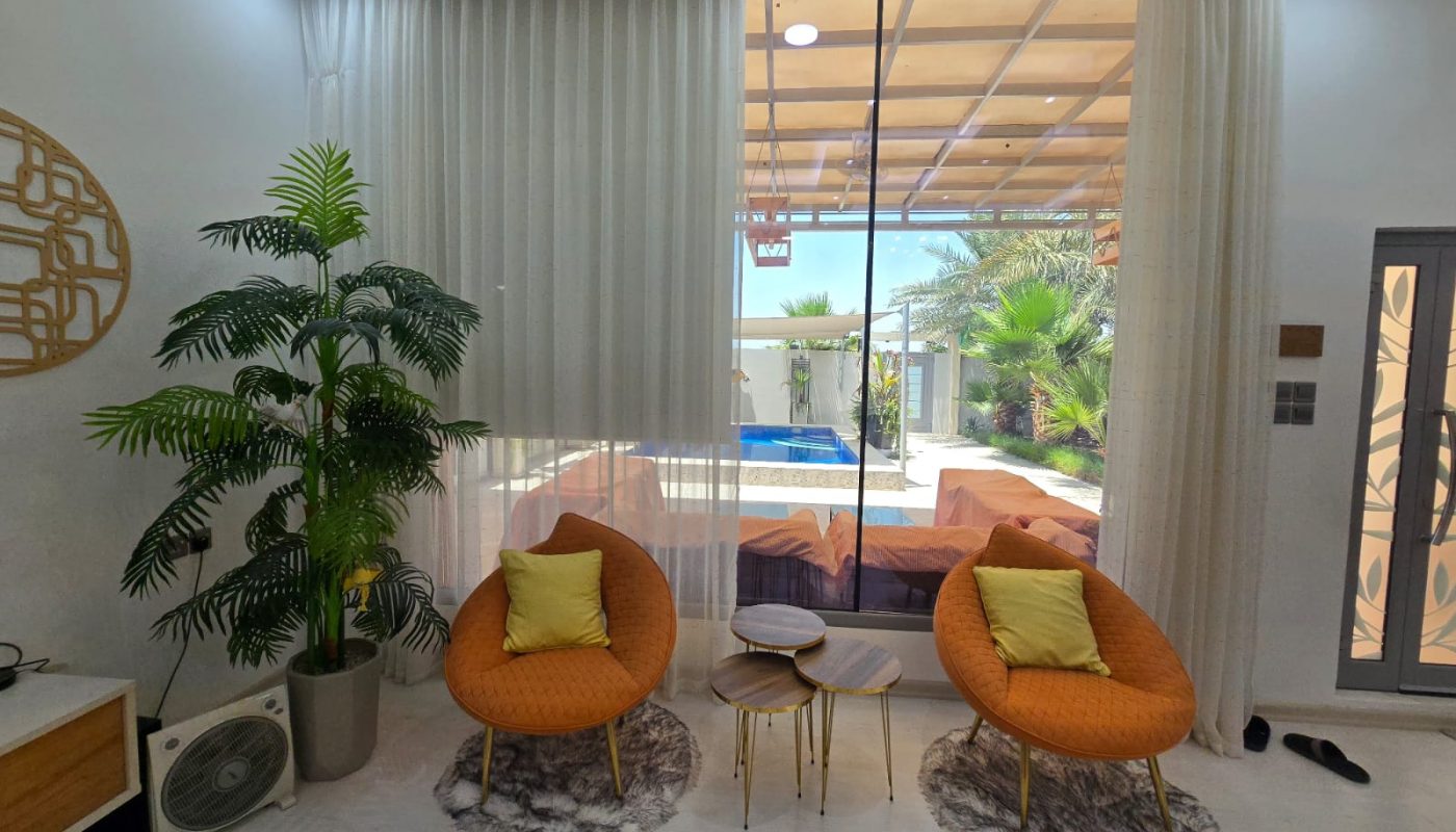 Two orange chairs with yellow pillows face a glass door overlooking a patio and pool, with a potted plant and small tables beside them in a modern living room.