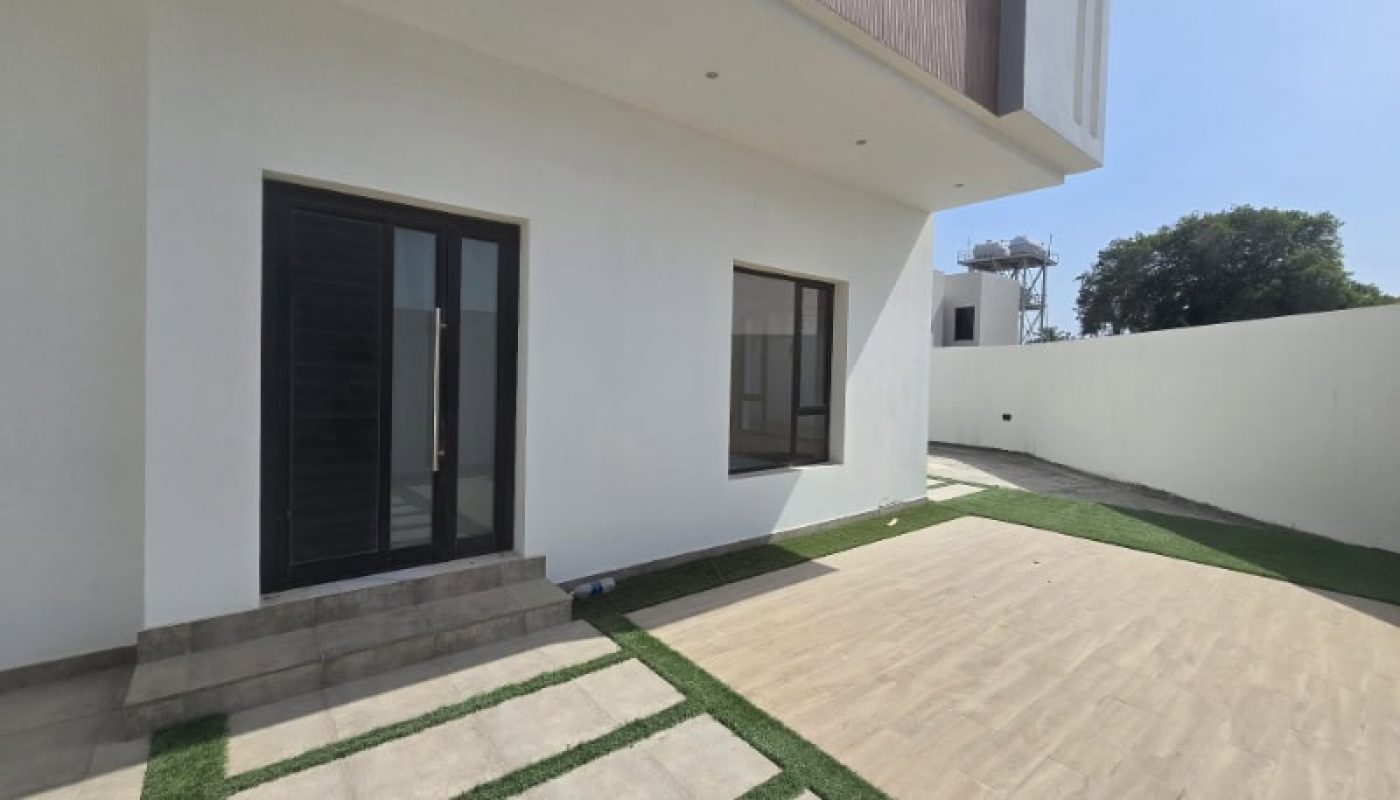 Modern house exterior with white walls, large glass doors, a window, tiled steps, artificial grass strips, and a paved courtyard under clear daylight.