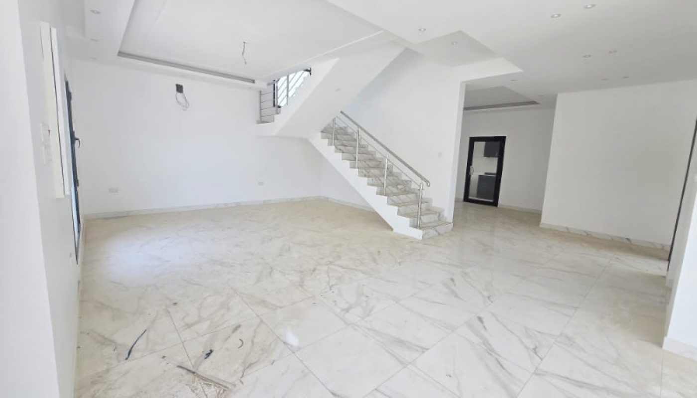Spacious, empty room with white walls, tiled floor, staircase with metal railing, and ceiling lights.