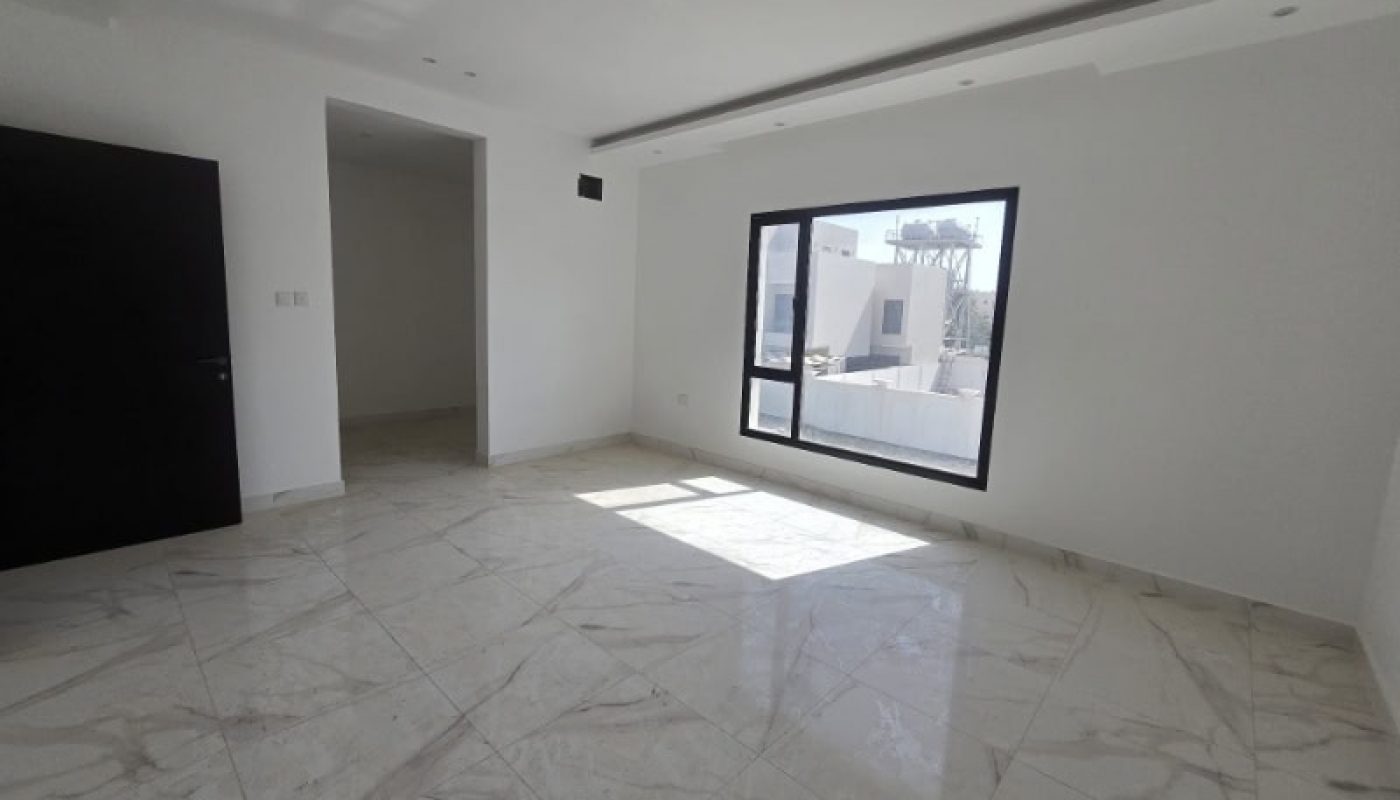 A bright, empty room with white walls, large window, marble-patterned tile floor, and an open doorway leading to another area.