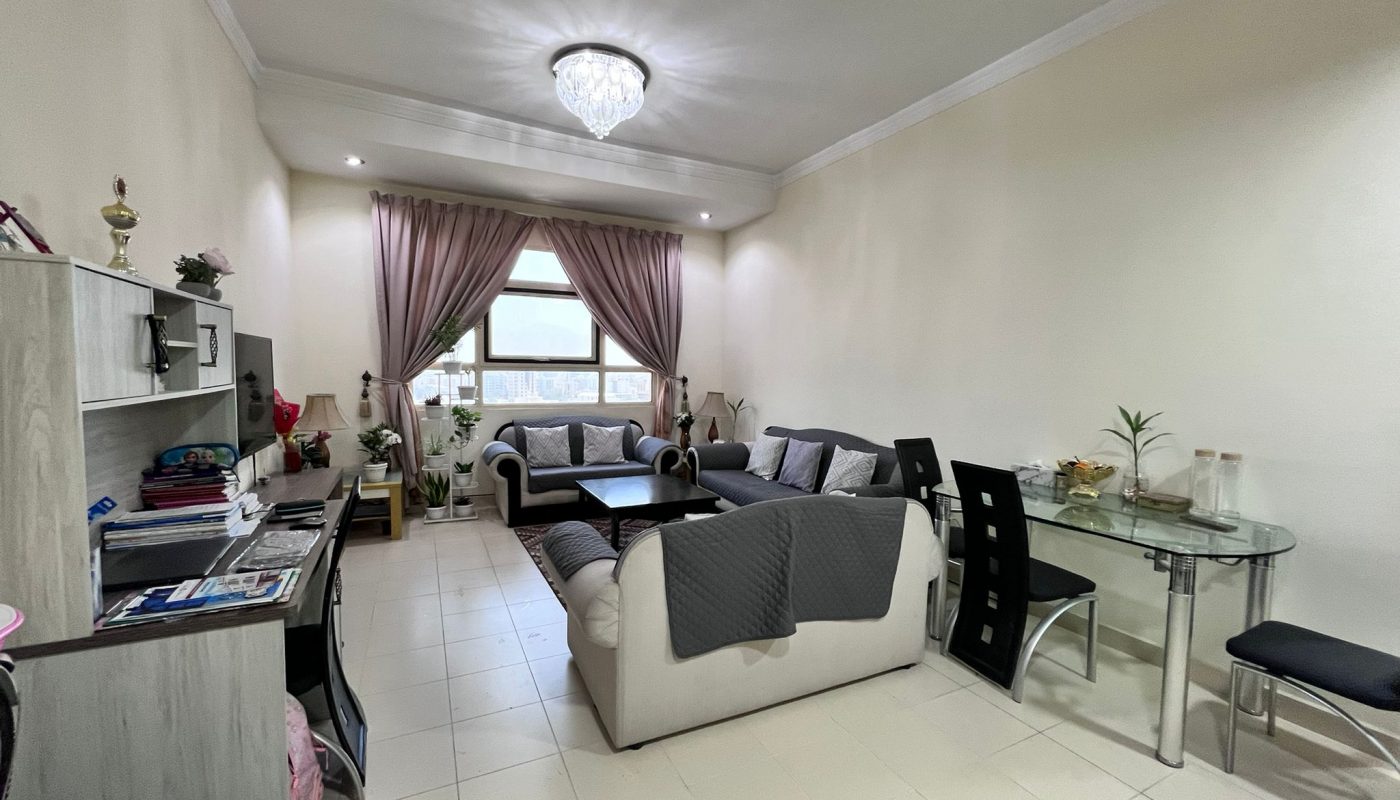 A living room with two sofas, a glass dining table with black chairs, a TV stand, shelves, and a window with curtains letting in natural light.
