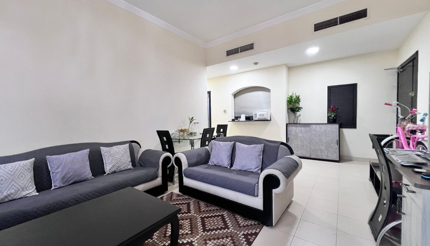 A modern living and dining area with two sofas, a coffee table, dining table with four chairs, and a kitchen in the background. The room features neutral colors and tiled flooring.