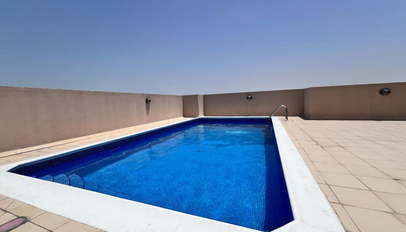 Rectangular outdoor swimming pool with clear blue water, surrounded by beige tiled flooring and high beige walls under a clear blue sky.