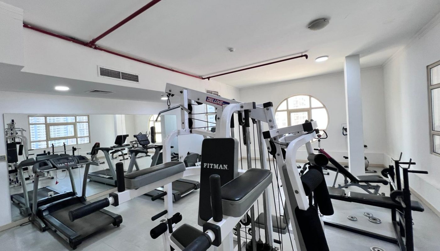 A gym with various exercise equipment, including weight machines, benches, free weights, and mirrors, in a well-lit room with white walls and large windows.