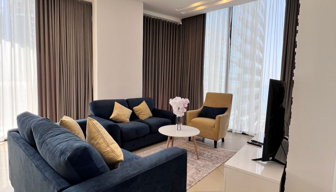 Modern living room with two blue sofas, a yellow armchair, a round coffee table, a TV on a white stand, and large windows covered by floor-length curtains.