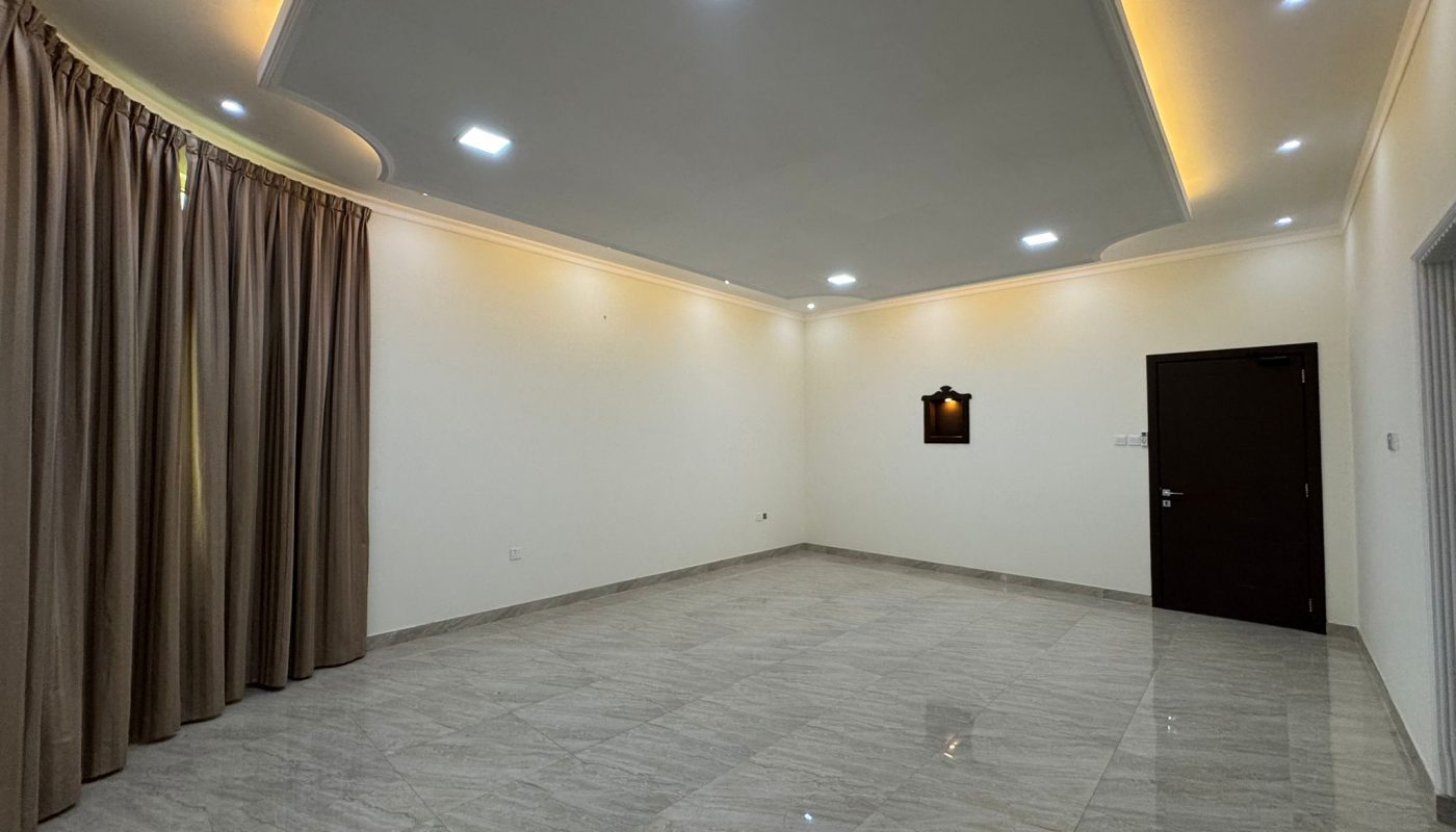 Empty room with tiled floor, cream-colored walls, ceiling lights, brown curtains on one side, a dark wooden door, and a small wall-mounted shelf with a light.
