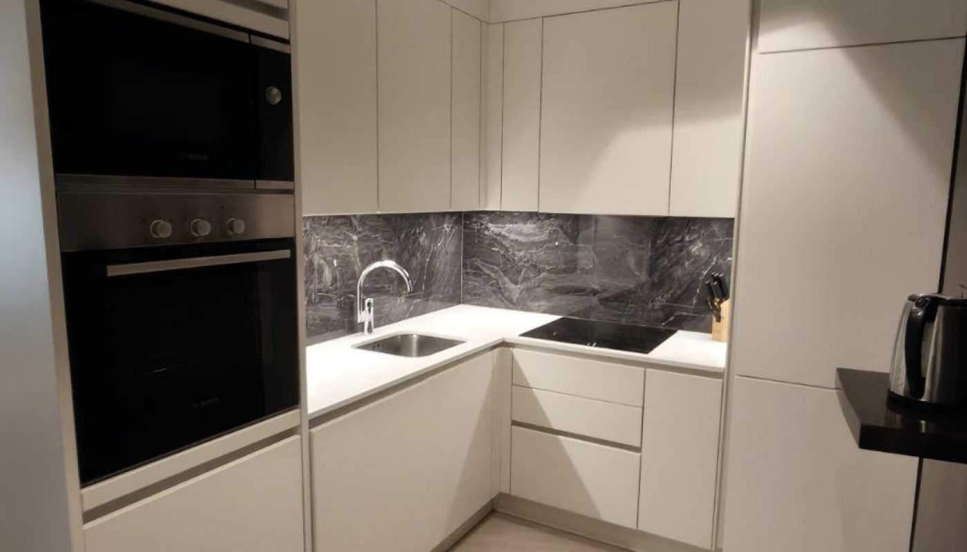 Modern kitchen with white cabinets, built-in appliances, under-cabinet lighting, marble backsplash, and a corner sink. The floor is tiled, and the overall design is minimalistic.