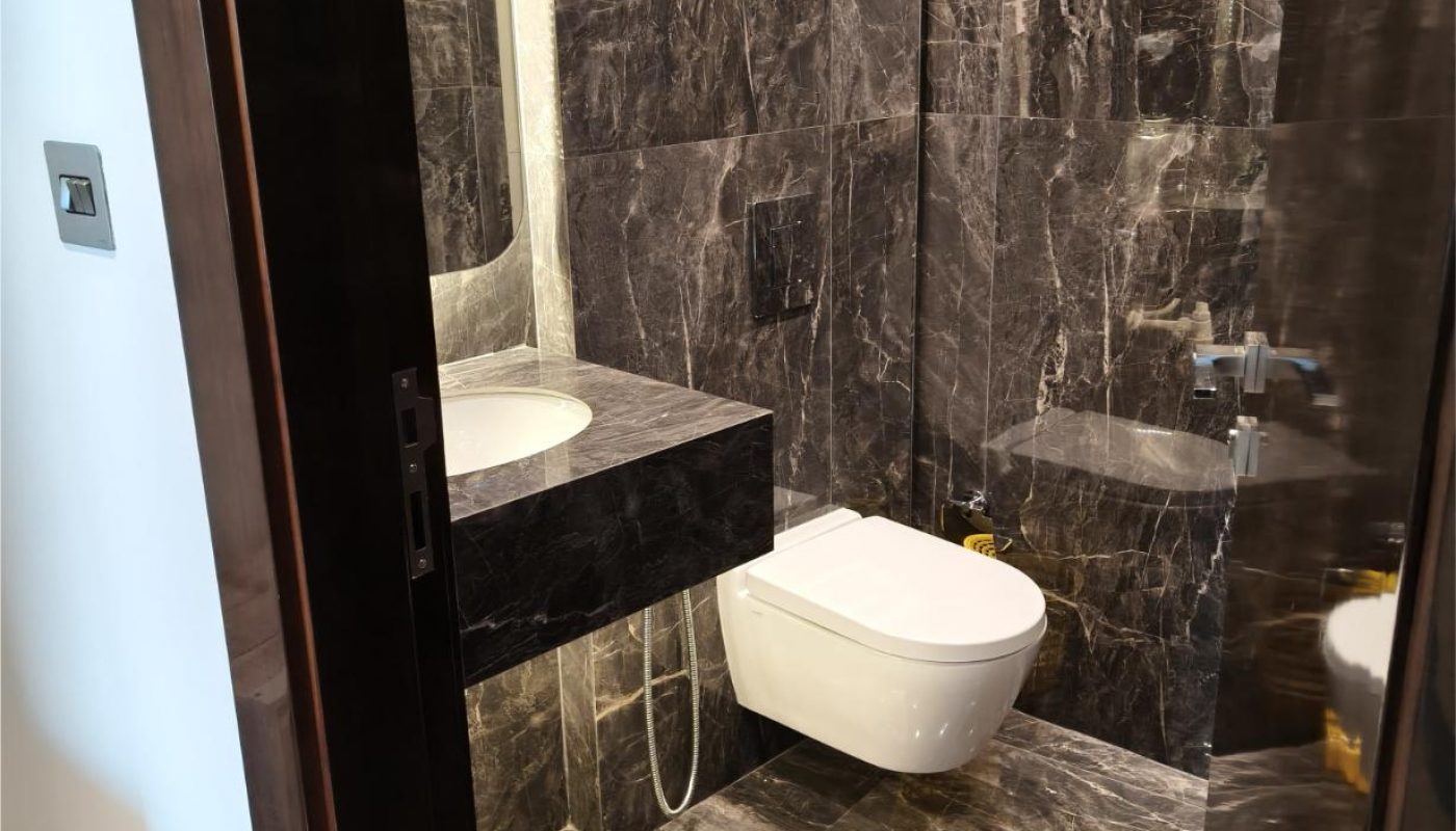 Modern bathroom with dark marble walls and floor, a wall-mounted toilet, a floating sink, and a large mirror. The door is open, and lighting is bright.