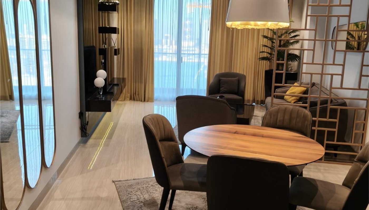 Modern living and dining area with round wooden table, four chairs, pendant light, sofa, large mirrors, sheer curtains, and decorative wooden divider. Marble floor and neutral tones throughout.