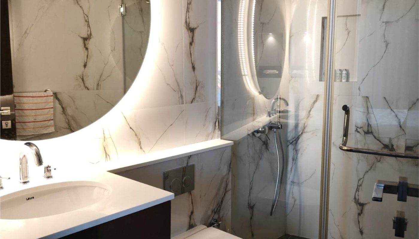 Modern bathroom with marble walls, round backlit mirror above a sink, wall-mounted toilet, and glass-enclosed shower with handheld showerhead.