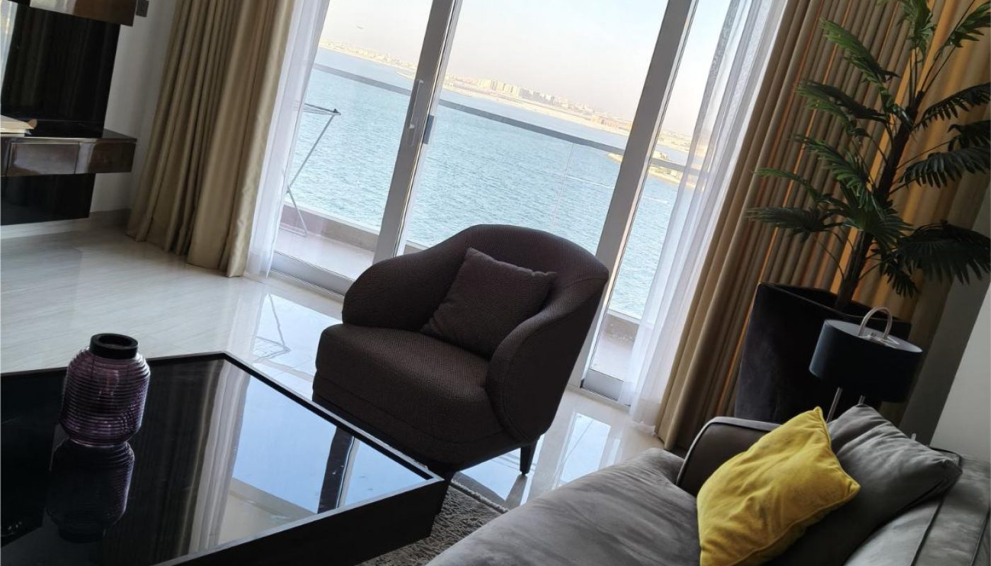 Modern living room with a gray sofa, yellow pillows, armchair, glass coffee table, large windows, beige curtains, and a view of water and distant buildings.