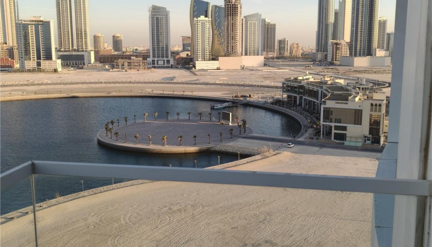 Cityscape view from a balcony showing high-rise buildings, a circular waterfront walkway, sand, and a calm body of water under a clear sky.
