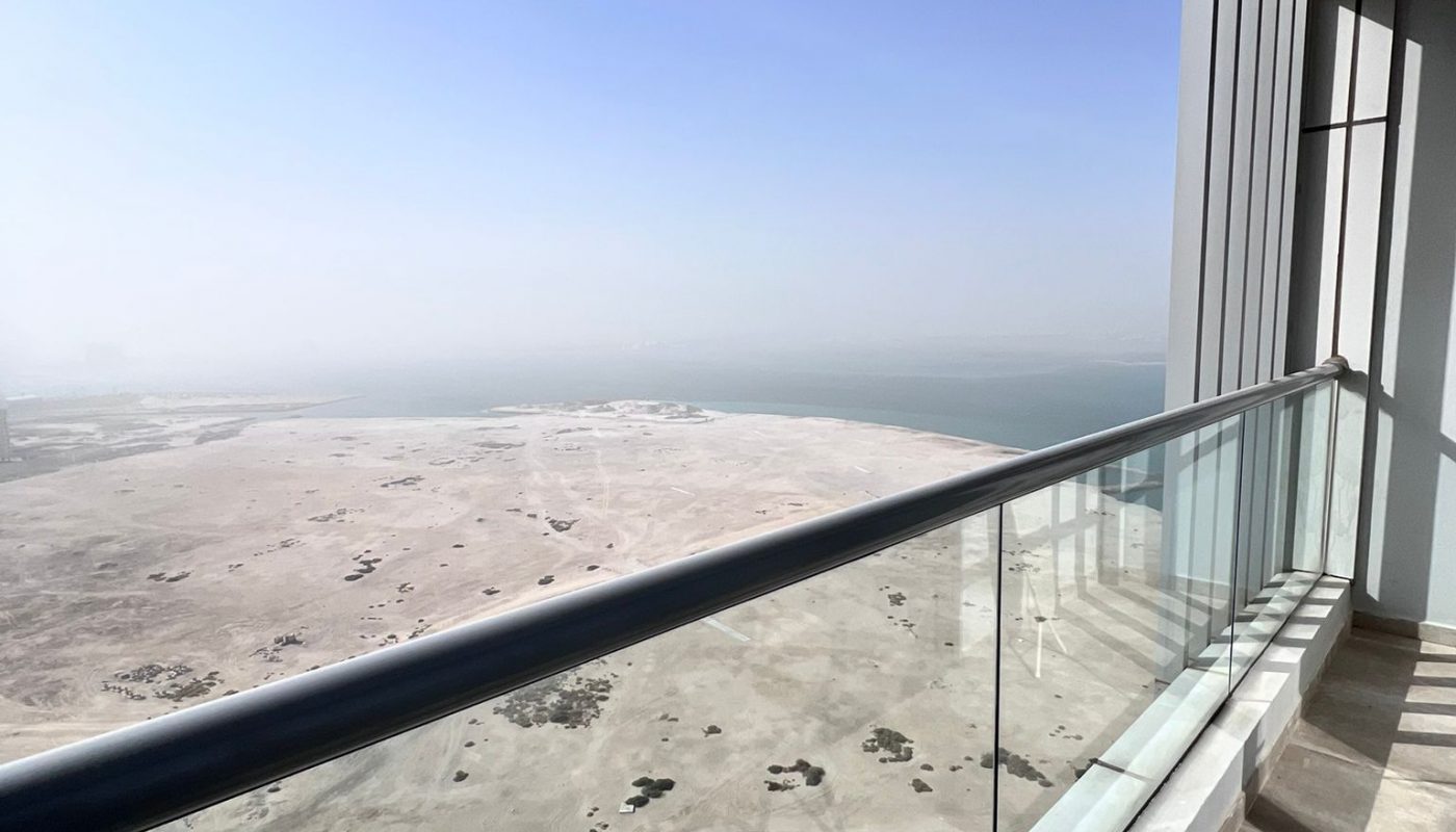 A high-rise balcony with glass railing overlooks a sandy, desert landscape and a distant body of water under a clear blue sky.