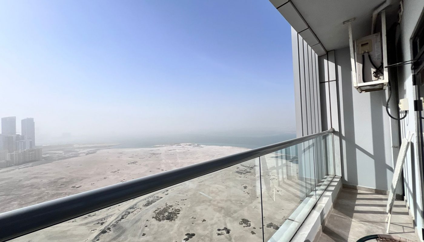 View from a high-rise balcony showing a sandy, undeveloped landscape, distant buildings on the left, and a large body of water under a clear blue sky.
