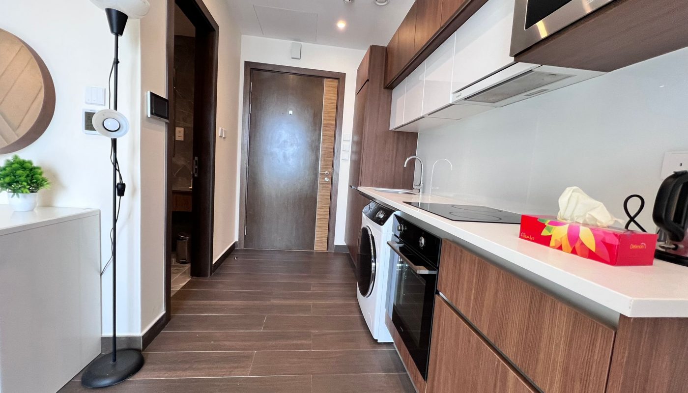 Modern kitchen with wood cabinets, white countertops, built-in appliances, washing machine, and a floor lamp next to a small table with a plant and mirror.