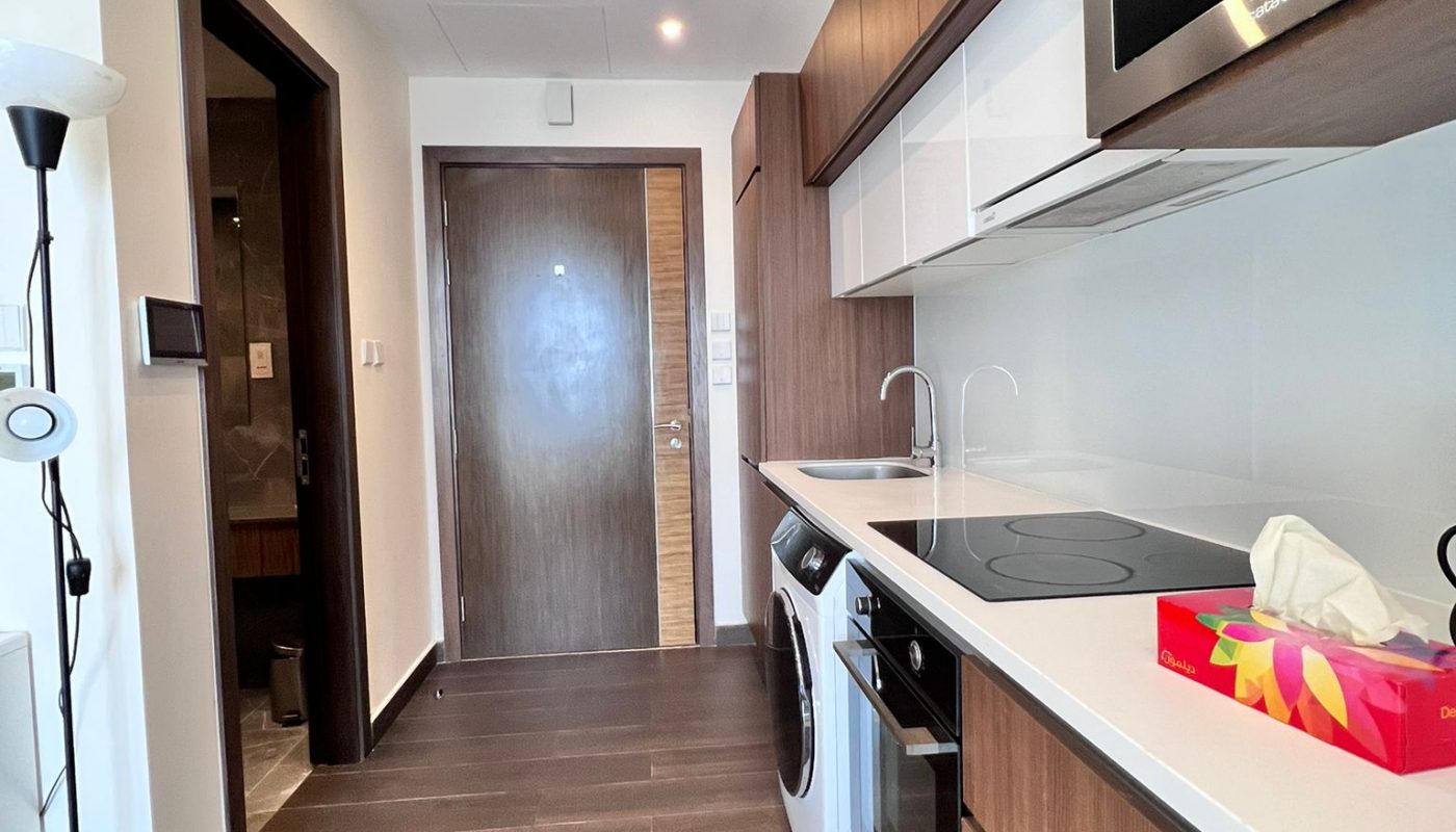 Modern kitchenette with wood cabinets, induction cooktop, microwave, washing machine, and a tissue box on the counter; a floor lamp and a door are visible in the background.