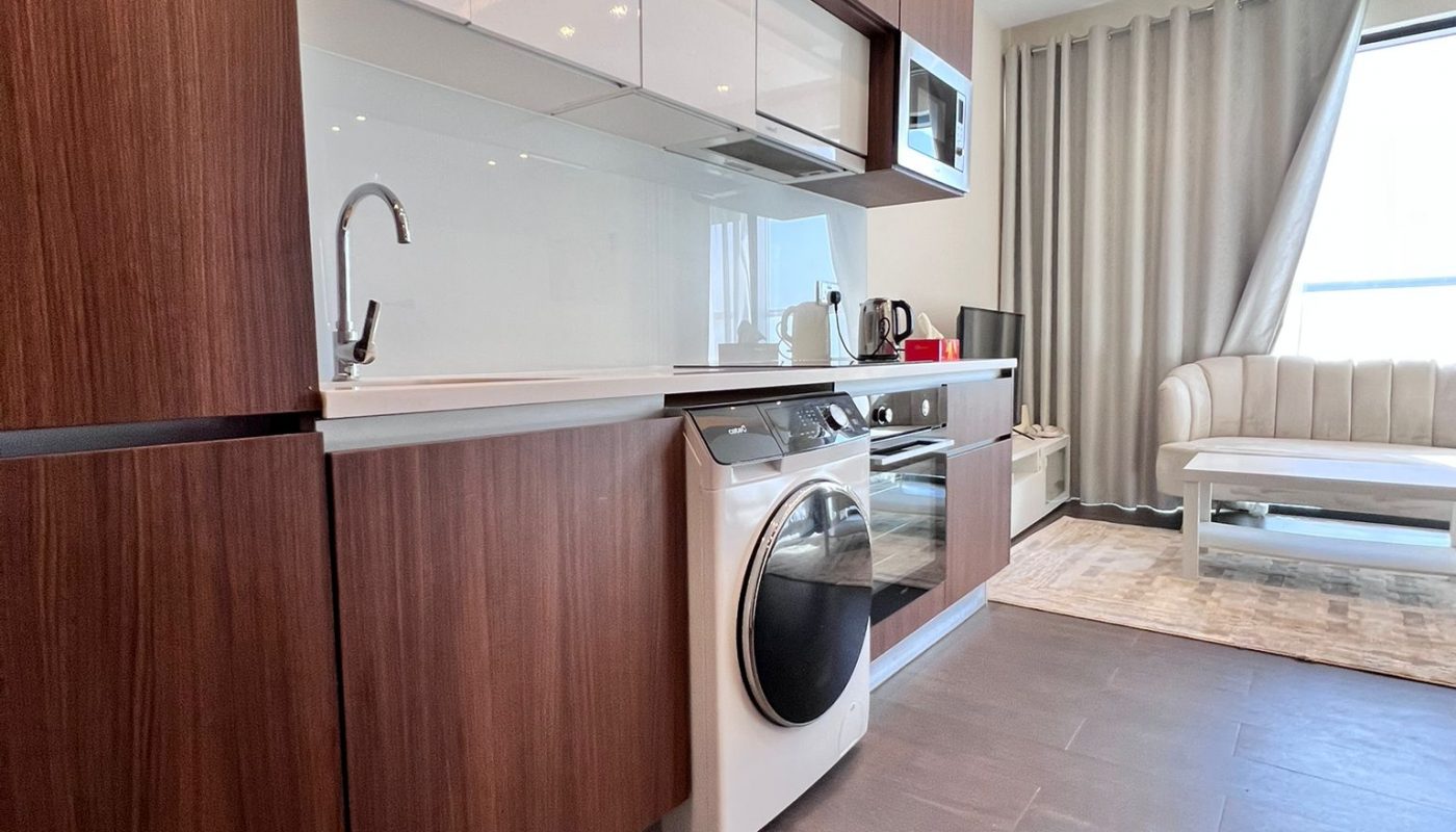 Modern kitchen with brown cabinets, white countertops, a washing machine, and a living area with a white sofa and large window with curtains.