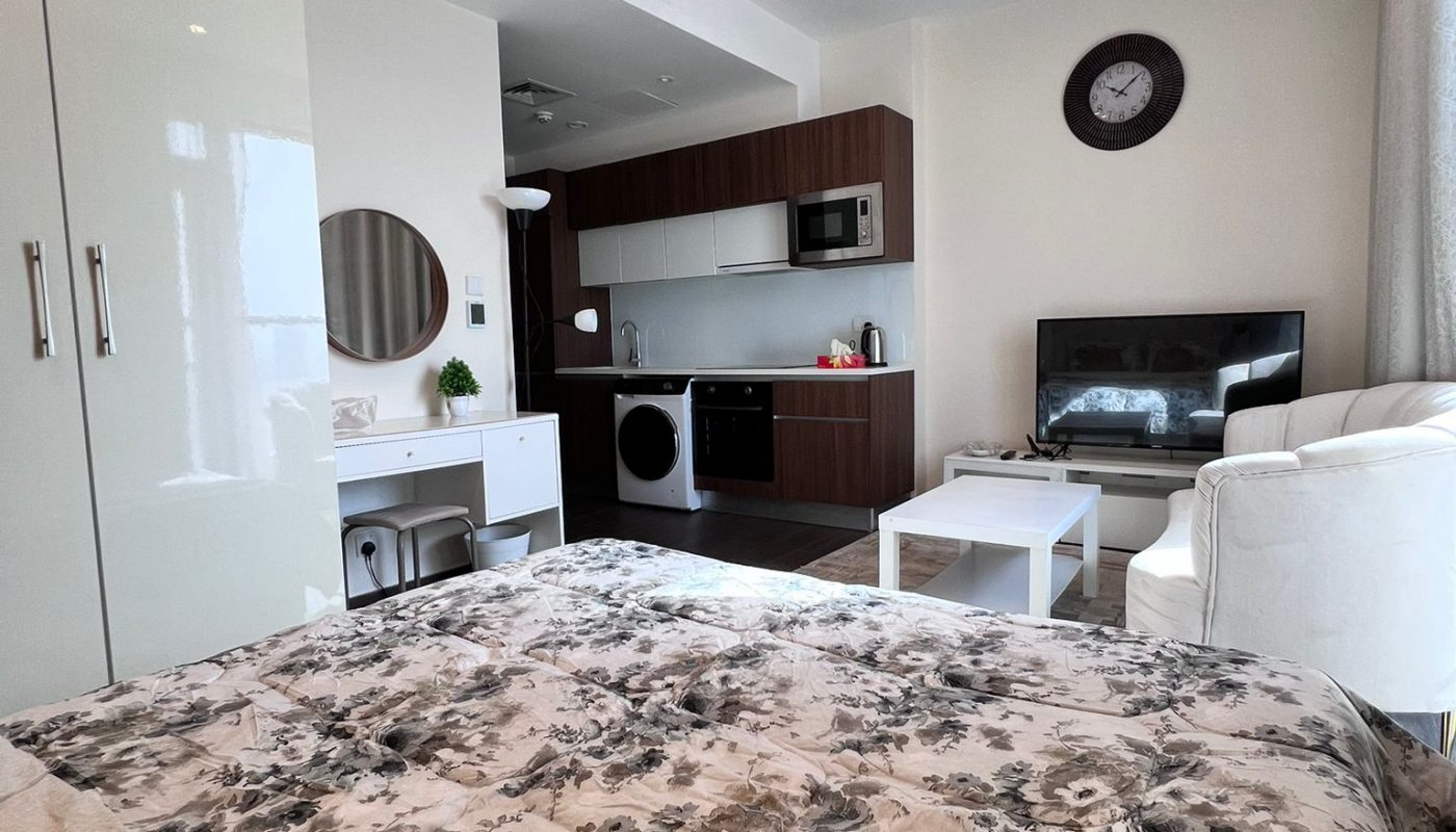 Modern studio apartment with a bed, kitchenette, washing machine, wall clock, TV, white armchair, and a small coffee table. Decor is neutral with floral bedding and light walls.