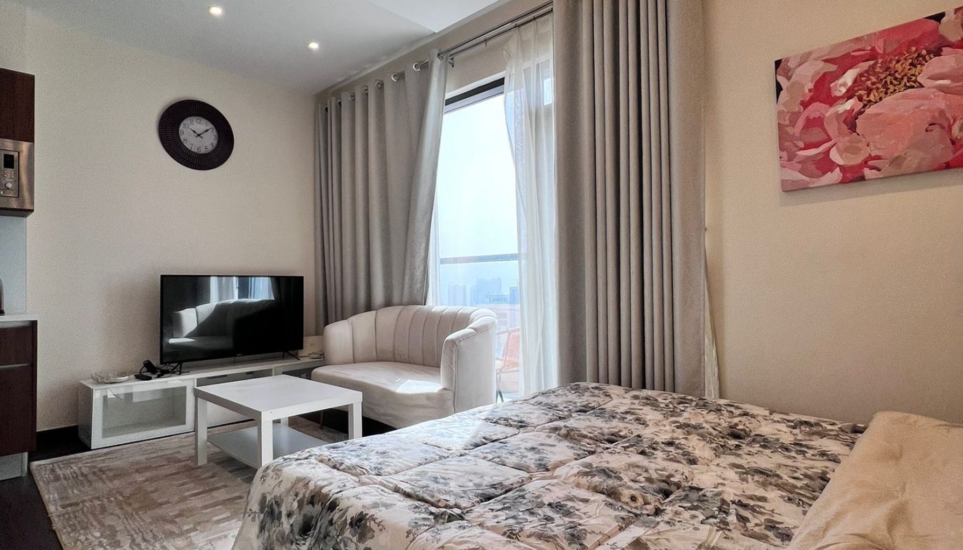 A modern studio apartment with a bed, sofa, coffee table, TV, wall clock, floral artwork, and large window with curtains letting in natural light.