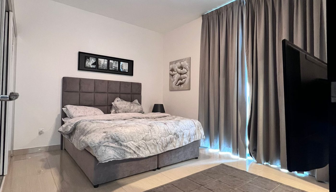 A modern bedroom with a gray upholstered bed, matching bedding, gray curtains, wall art above the bed, a nightstand with a lamp, a gray rug, and a wall-mounted TV.