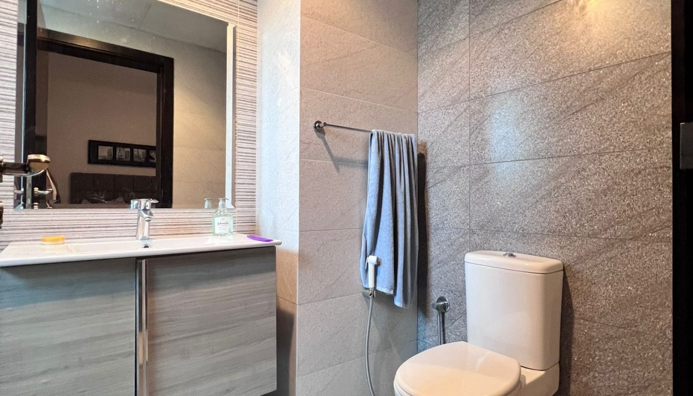 Modern bathroom with gray tiled walls and floor, featuring a toilet, a sink with cabinets, a mirror, and a towel hanging on a rail.