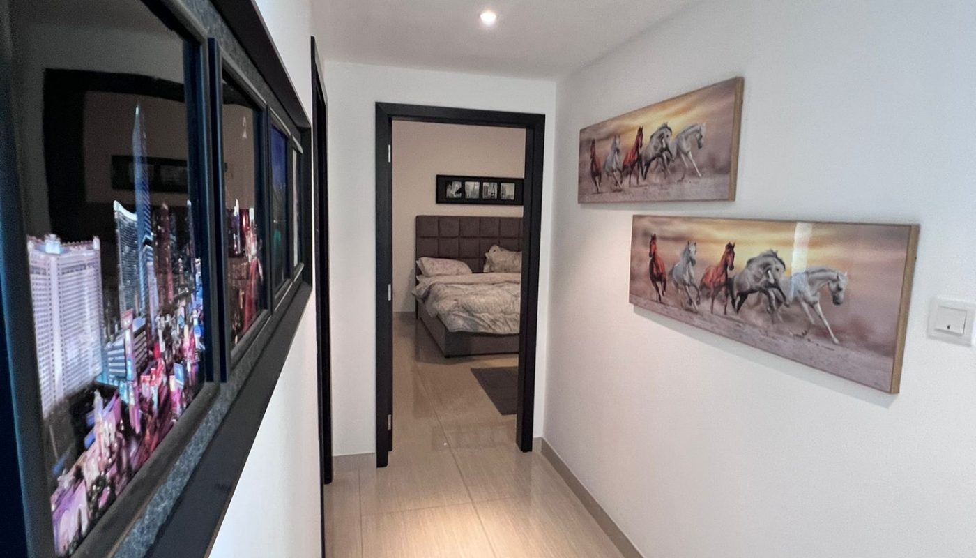 A hallway with framed pictures on the walls leads to a bedroom with a neatly made bed and a headboard.