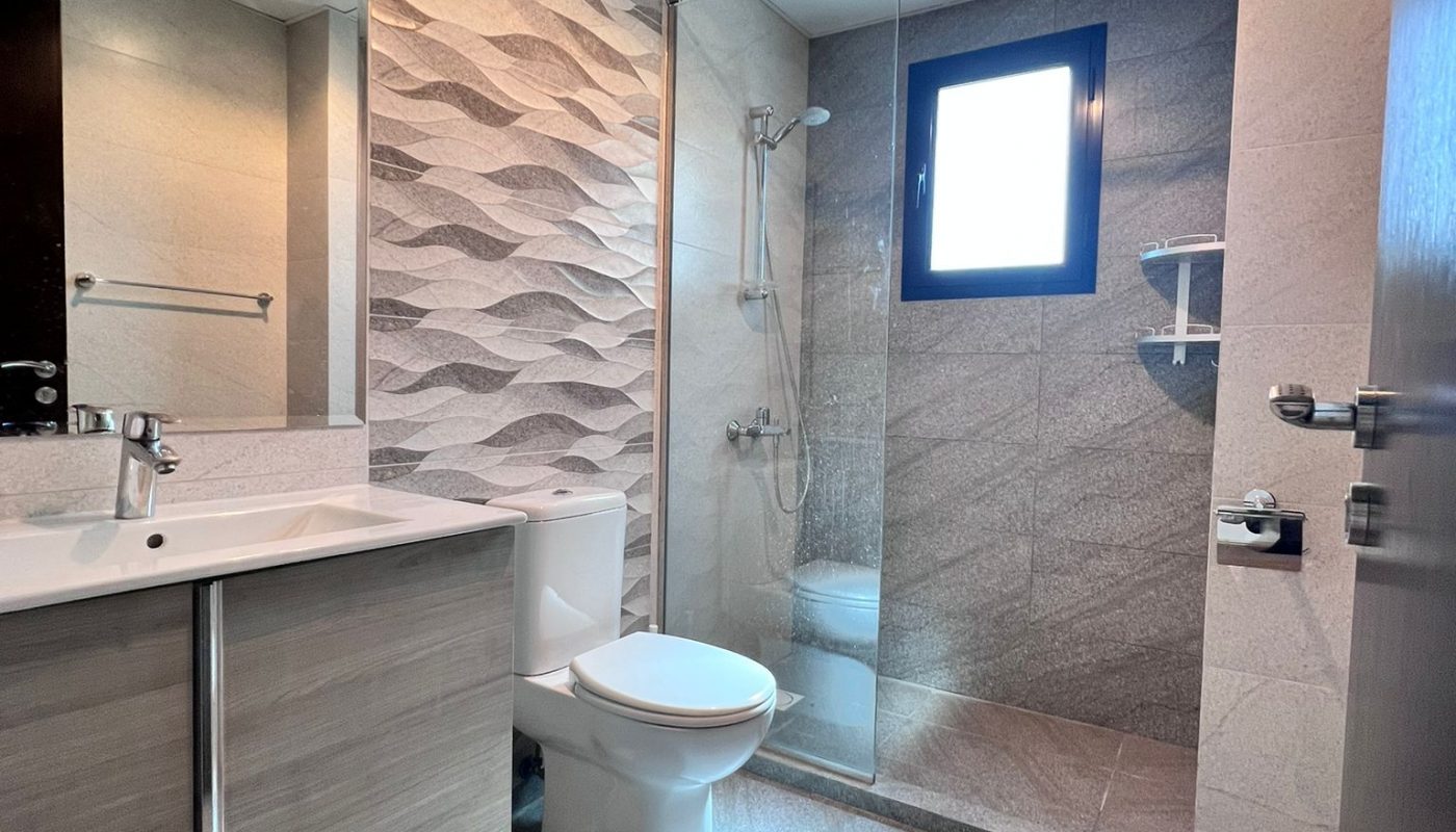 Modern bathroom with a sink, toilet, and glass-enclosed shower; features textured wall tiles and a window above the shower area.
