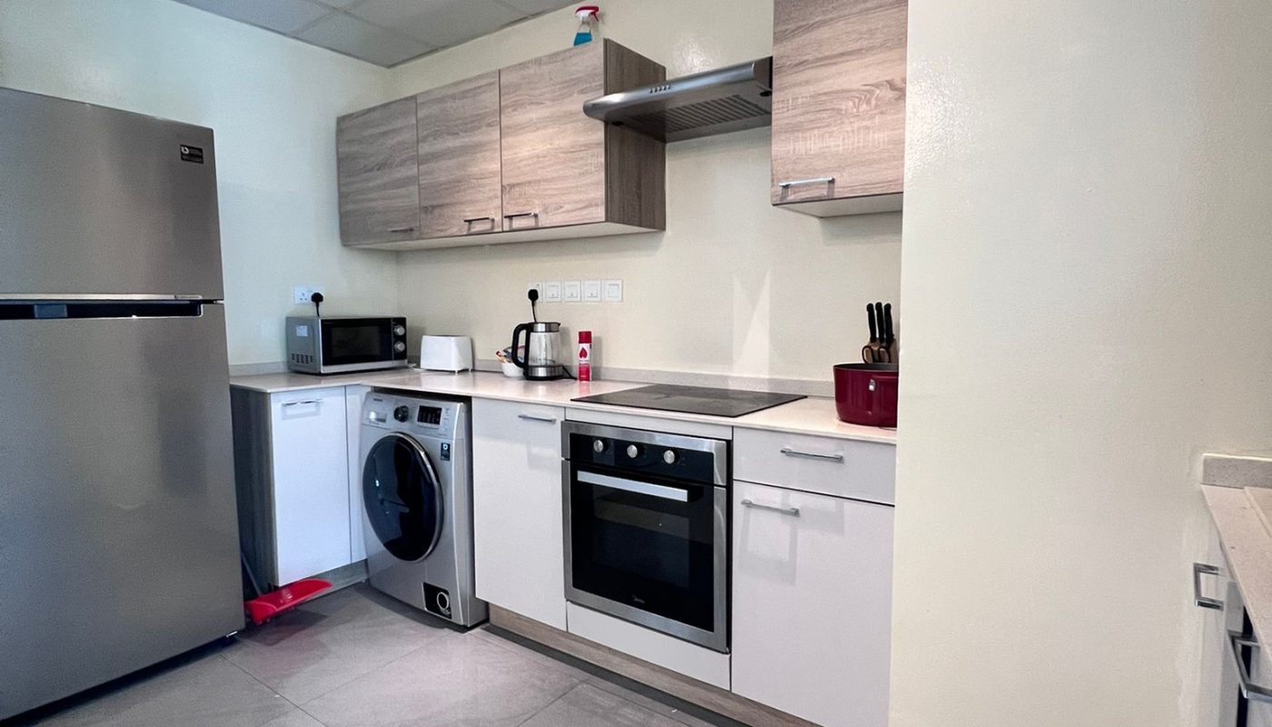Modern kitchen with grey tile floor, stainless steel refrigerator, oven, washing machine, microwave, light wood cabinets, and utensils on the counter.