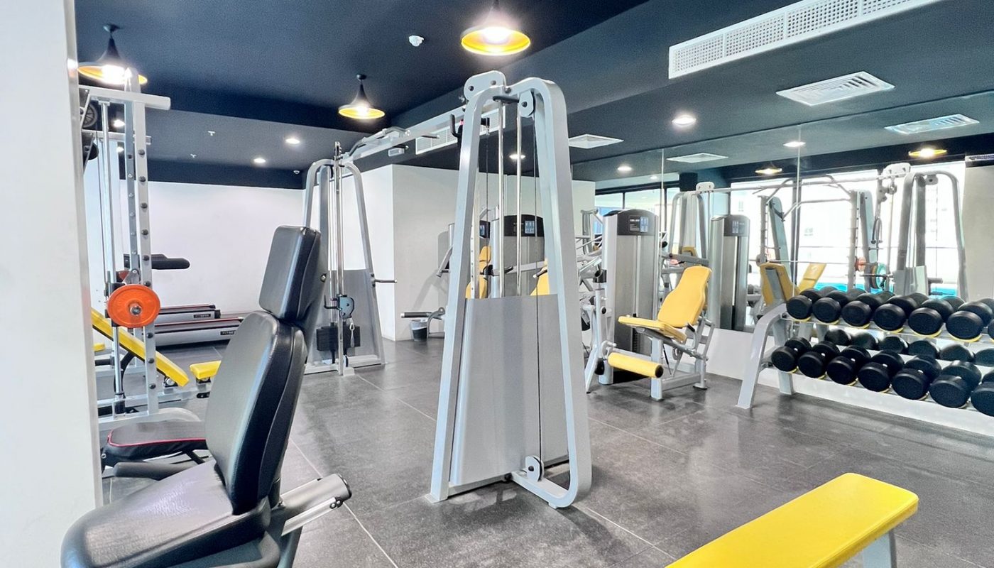 Modern gym interior with various exercise machines, yellow benches, free weights, and large mirrors on the walls under black ceiling with hanging lights.