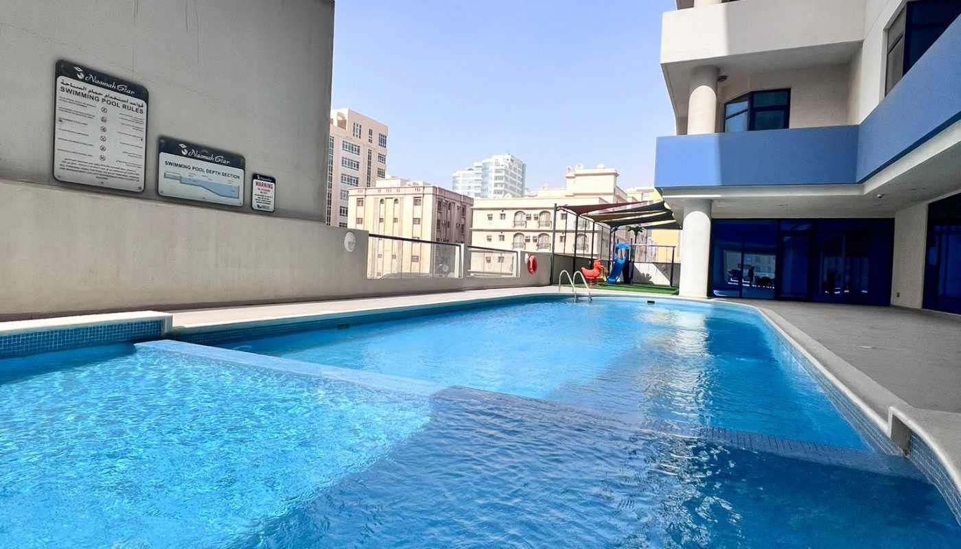 Outdoor swimming pool beside a modern multi-story building under a clear blue sky, with city buildings in the background.