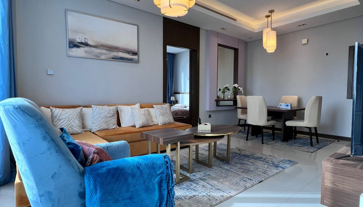 A modern living room with a blue armchair, beige sofa, wooden coffee tables, dining area with white chairs, and contemporary lighting fixtures.