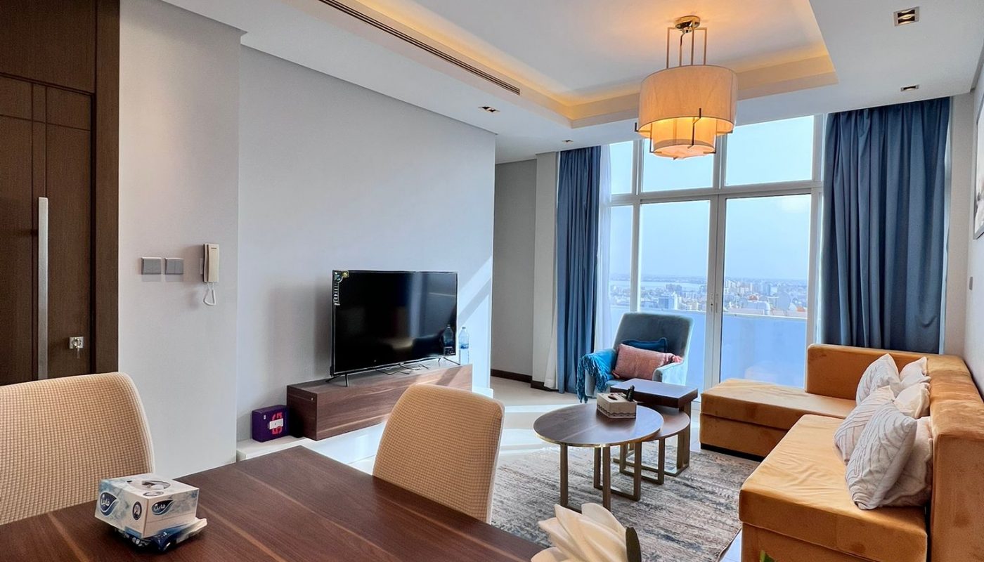 Modern living and dining area with a wooden table, beige chairs, yellow sofa, flat-screen TV, large windows, and blue curtains, featuring bright overhead lighting.