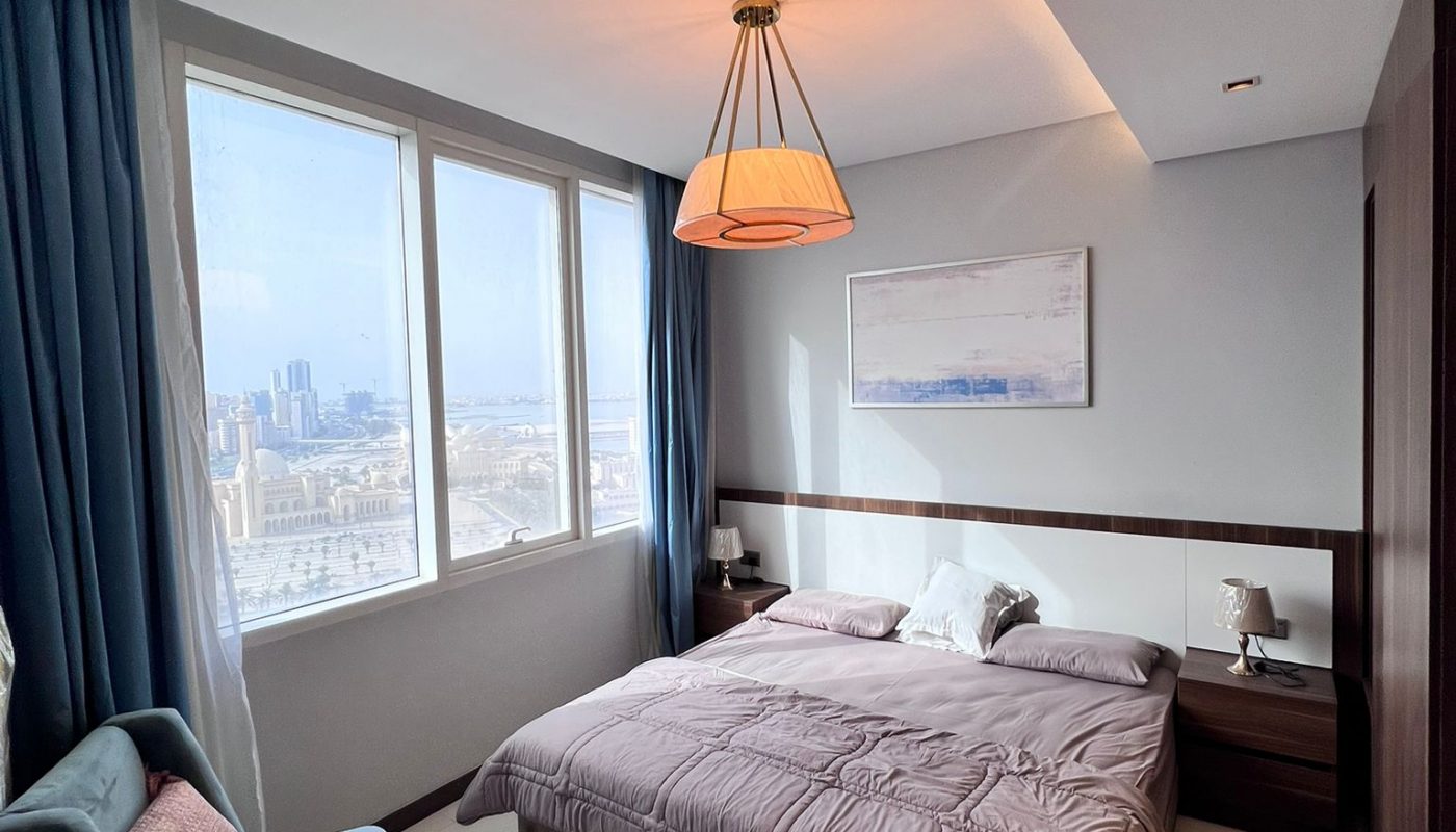 Modern bedroom with a large window, city view, double bed with mauve bedding, blue curtains, small velvet chair, side tables, and wall art above the bed.