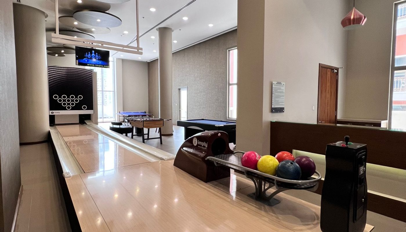 A modern indoor bowling alley with a polished lane, colorful bowling balls, a scorekeeping console, and game tables in the background.