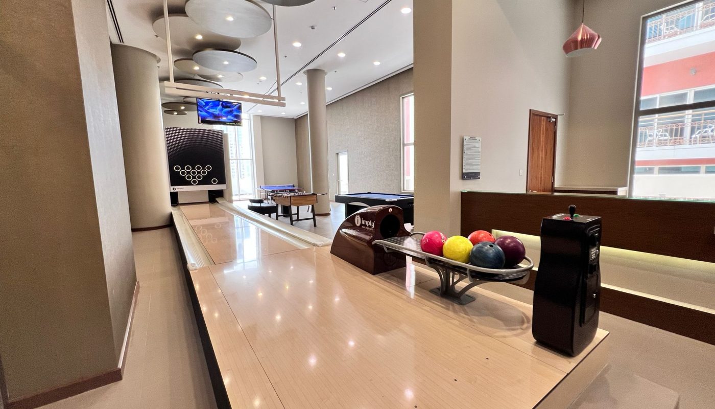 A modern indoor recreation room with a bowling lane, colorful bowling balls, a pool table, foosball table, and a television mounted on the wall.