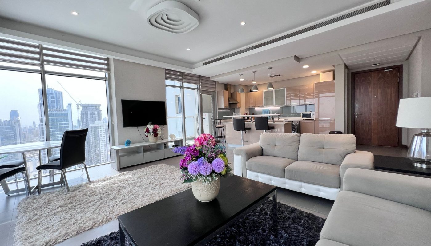 Modern living room with a beige sofa, black coffee table with a flower arrangement, TV on the wall, open kitchen, and large windows overlooking a city skyline.