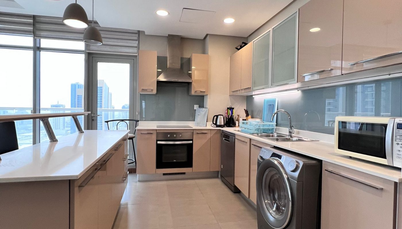 Modern kitchen with beige cabinets, built-in oven, washing machine, microwave, sink, and hanging pendant lights. Large windows provide natural light and a city view.