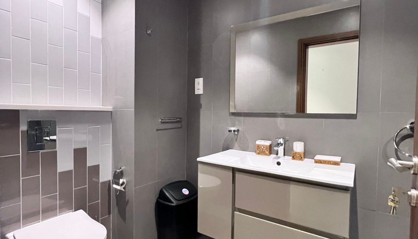 Modern bathroom with gray tiles, wall-mounted toilet, floating vanity with sink, large mirror, black trash bin, and assorted toiletries on the counter.