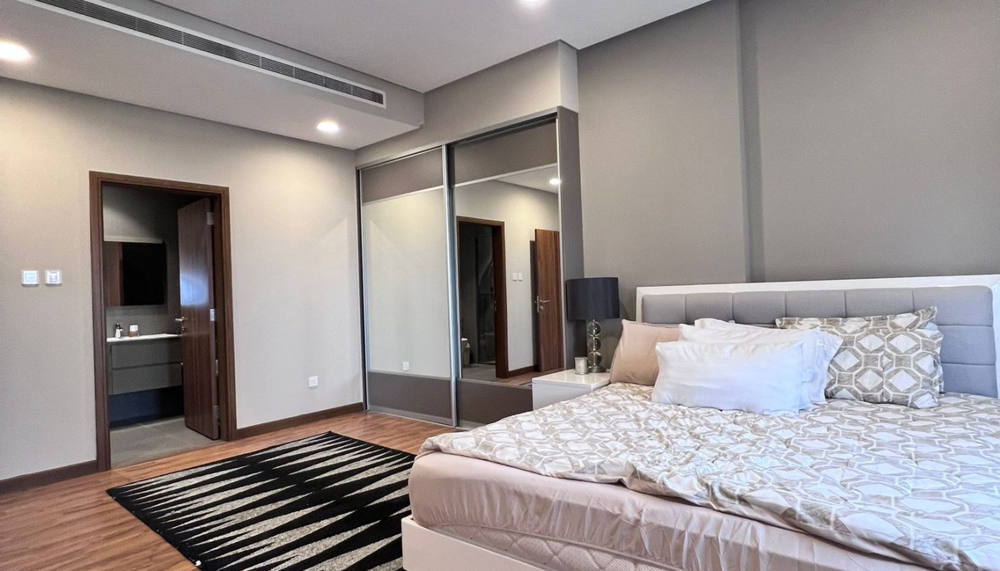A modern bedroom with a white bed, patterned pillows, a black lamp, a striped rug, large mirrored closet doors, and wood flooring. An open door leads to a bathroom.