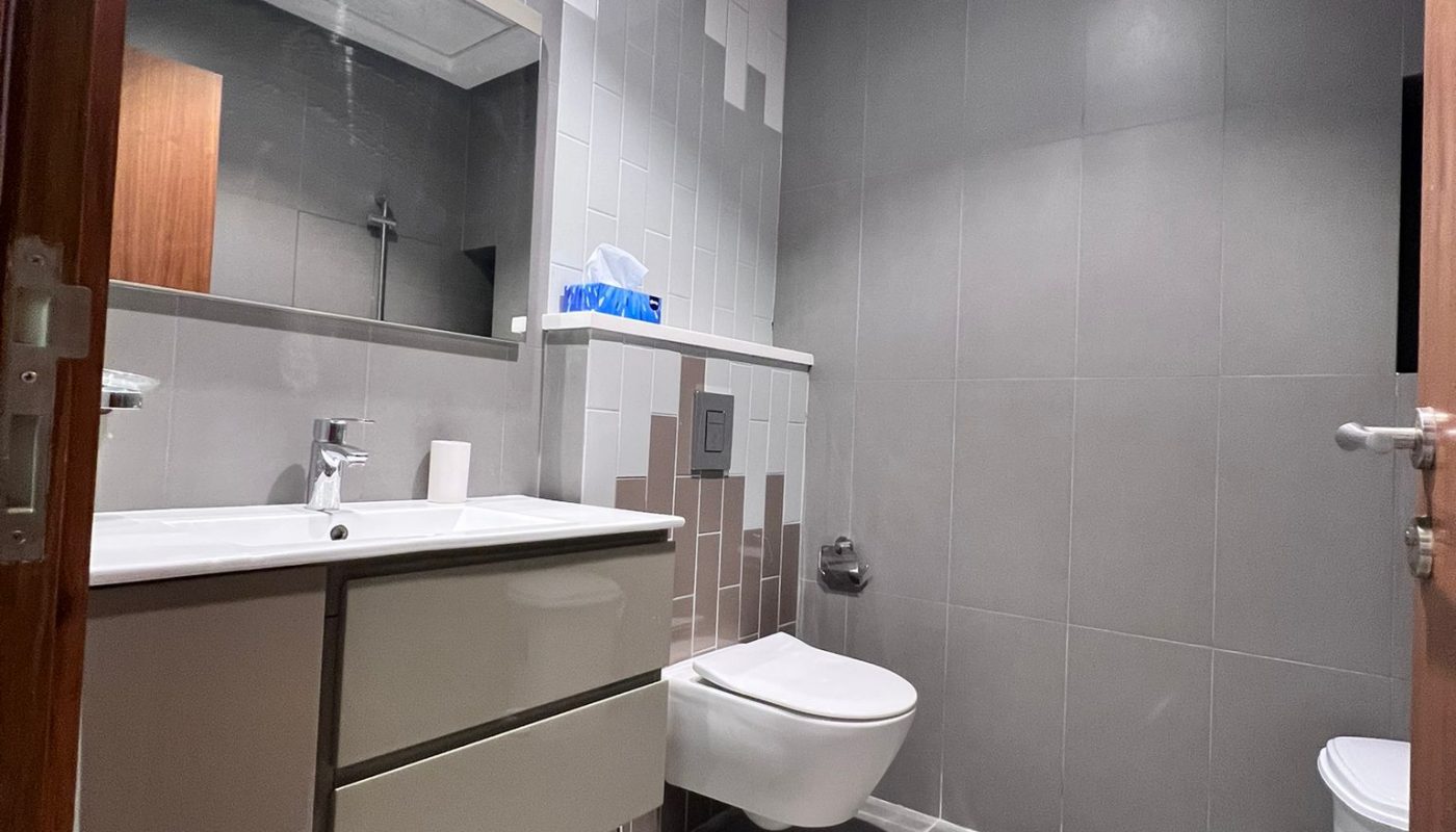 Modern bathroom with gray tiles, wall-mounted sink and cabinet, floating toilet, tissue box on the counter, and large mirror under recessed ceiling lights.