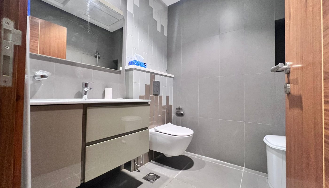 Modern bathroom with gray tiles, wall-mounted toilet, floating vanity with sink, mirror, and a small trash bin beside the toilet.