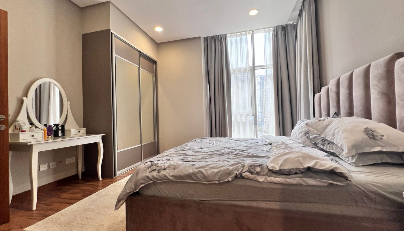 A modern bedroom with a large bed, a vanity table with a round mirror, a wardrobe, wood flooring, and natural light coming through a tall window with curtains.
