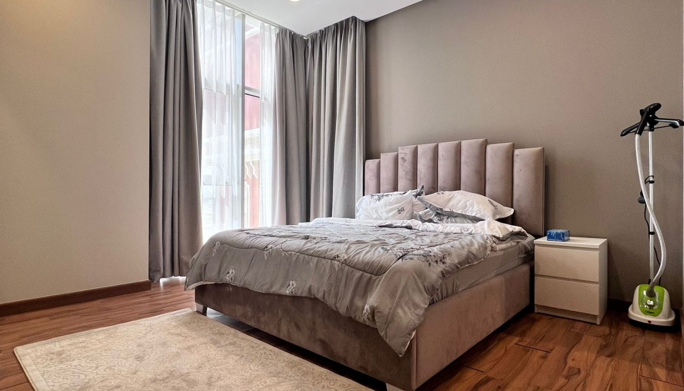 A modern bedroom with a double bed, beige headboard, nightstand, floor lamp, and a vacuum cleaner on wooden flooring with a light rug. Curtains cover tall windows.