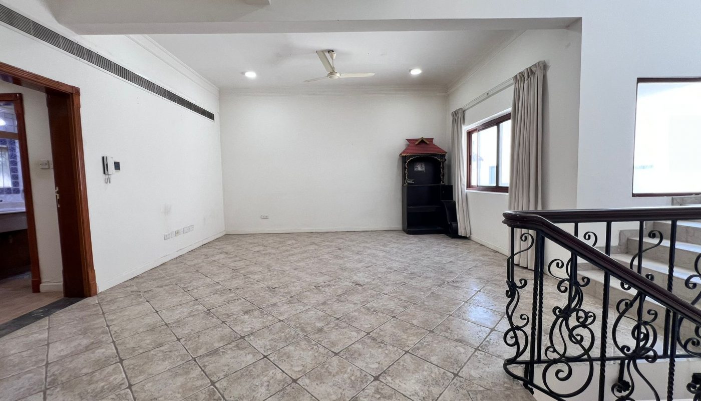 Empty tiled living room with white walls, ceiling fan, two windows with curtains, a small black cabinet in the corner, and decorative stair railing on the right.
