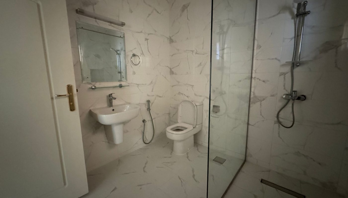 A bathroom with white marble-patterned walls and floor, featuring a toilet, wall-mounted sink, mirror, bidet spray, and a glass-enclosed shower area with a mounted showerhead.