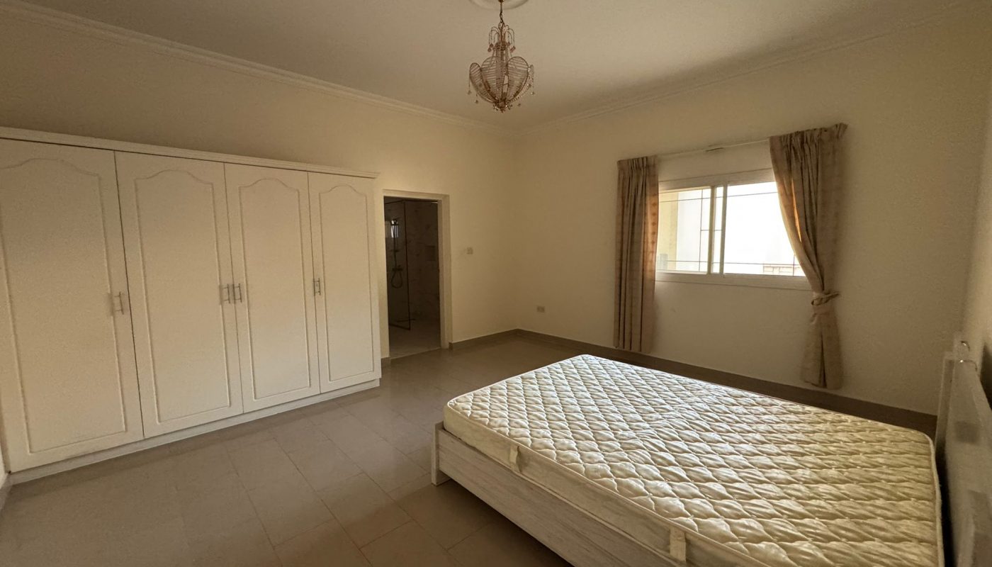 A sparsely furnished bedroom with a mattress on a bed frame, a large white wardrobe, a window with curtains, and a chandelier on the ceiling.