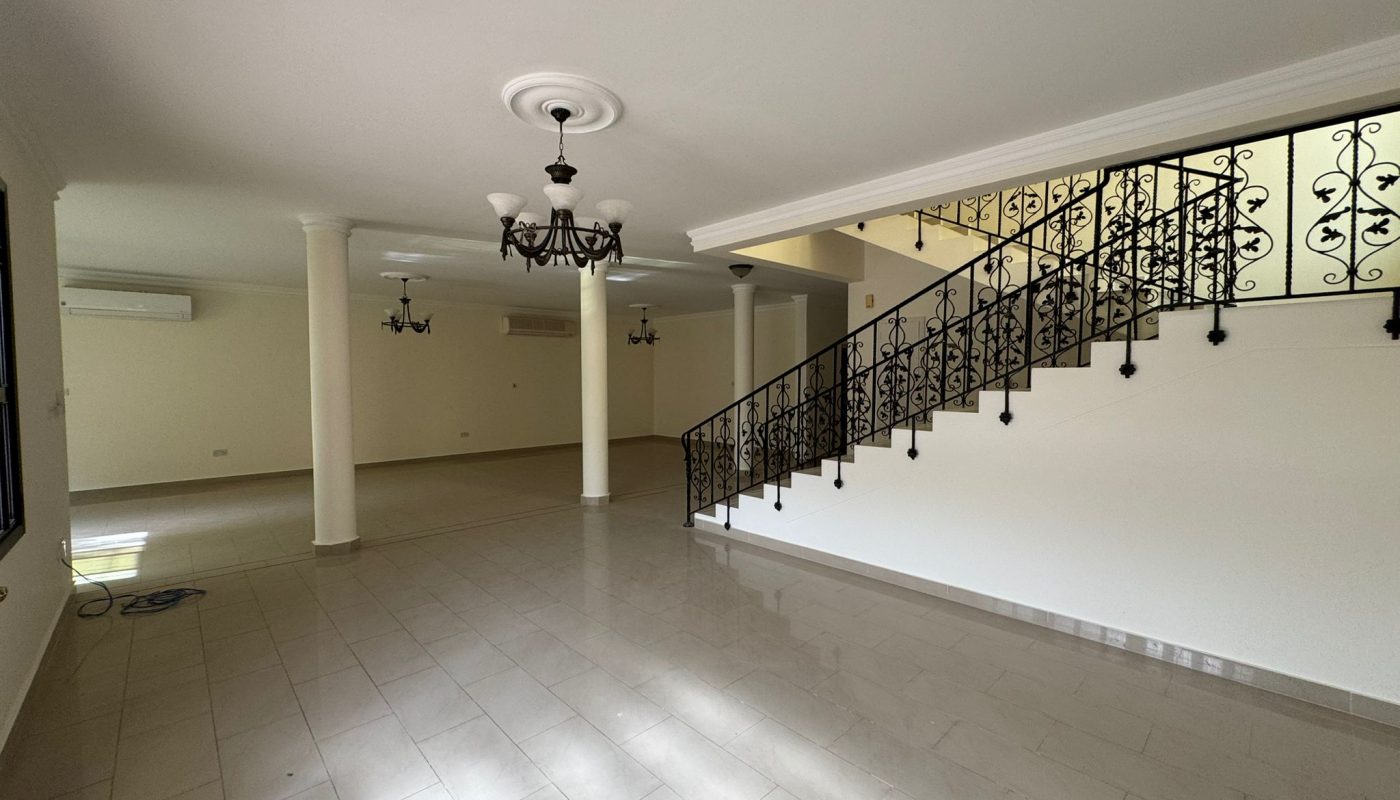 Spacious empty living room with tiled floor, white walls, two chandeliers, pillars, and an ornate black metal staircase railing.