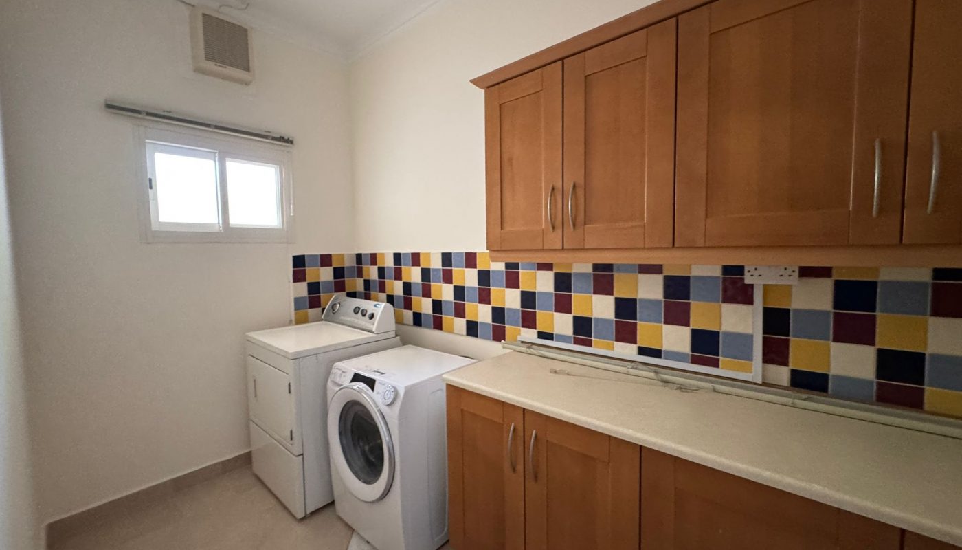 Laundry room with a washing machine, dryer, wooden cabinets, beige countertops, and a multicolored tiled backsplash under a window.