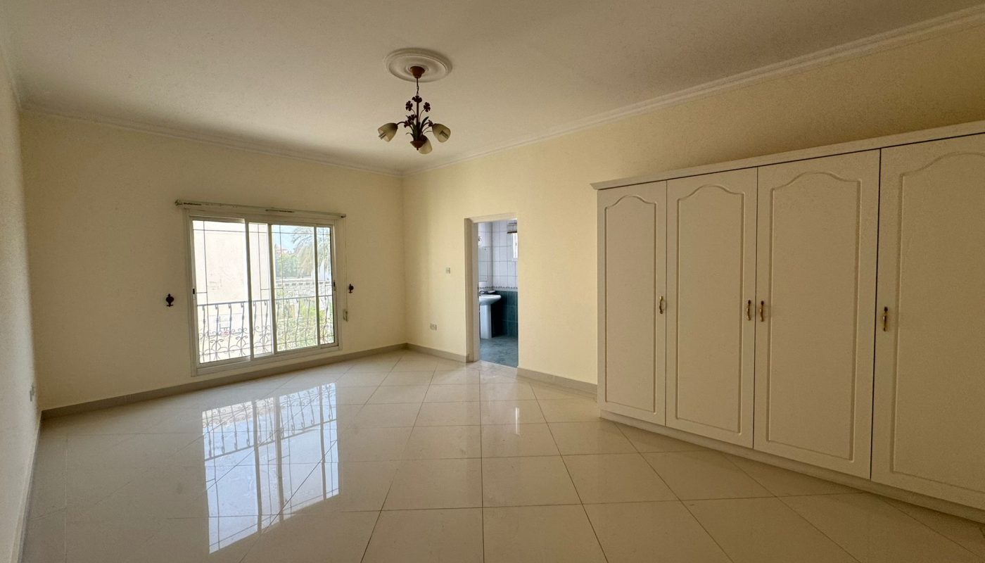 Empty room with tiled floor, built-in white wardrobe, window with blinds, overhead light fixture, and an open doorway leading to another area.