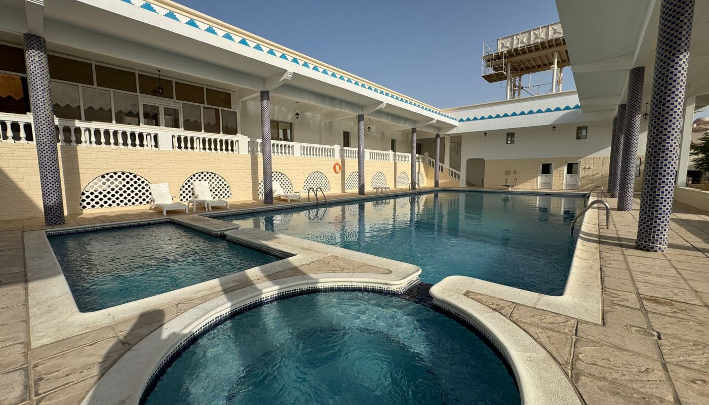 A large outdoor swimming pool with an attached circular hot tub, surrounded by tiled deck and covered walkways, under a clear sky.